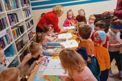 Dzieci stoją przy stolikach. Przed nimi leżą kartki z narysowanym "molem książkowym". Wśród nich pani w czerwonej bluzce, z boku pan w koszuli w kratkę. Na ścianie napis: czytelnia.