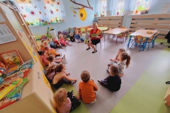 Dzieci siedzą na szarej podłodze w kole w sali przedszkolnej. Przy nich na krzesełku siedzi pani. W rękach trzyma książkę . W tle okno z kolorowymi kwiatami.