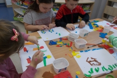 Przy stoliku przedszkolnym siedzi troje dzieci. Układają one na białych kartkach barwną kompozycję z kolorowych wycinków bibuły.. Na stoliku leżą pojemniki z wodą, pędzle, nożyczki i kawałki kolorowej bibuły. W tle widać szafki przedszkolne