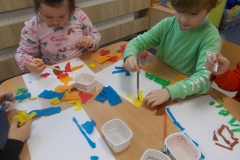 Przy stoliku przedszkolnym widać siedzącego chłopca w zielonej bluzie z białymi wstawkami na rękawach oraz dziewczynkę w różowej bluzie z nadrukiem. Za pomocą pędzelków pokrywają wodą kawałki kolorowej bibuły rozłożone na kartce. Widać również dłonie dwóch kolejnych dzieci siedzących przy tym stoliku. A na nim leżą pojemniki z wodą, pędzelki i skrawki kolorowej bibuły.