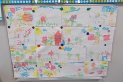 Wystawka prac dziecięcych. Na tablicy przypięte magnesami znajdują się kolorowe prace dzieci wykonane techniką ,, malowanie bibułą’’. Rozpoznać można kształty chmur, słońca, kwiatów, trawy.