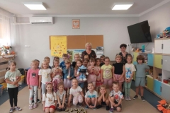 Grupa dzieci ustawiona w półkolu. Przed nimi kuca 6 dzieci. Na beżowej podłodze leży 10 pszczółek. 4 dzieci trzyma w rękach maskotki. Za dziećmi stoją 2 panie. W tle na ścianie wisi godło, tablica korkowa i z boku telewizor.