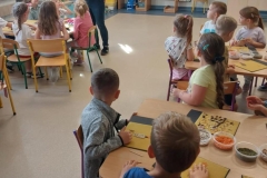 Dzieci siedzą przy stolikach. Na nich tabliczki z wosku pszczelego. W pojemnikach różnokolorowe ziarenka. Przed dziećmi stoi pani w ciemnych spodniach i kolorowej bluzce. W rękach trzyma maskotkę pszczoły.