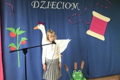 Zdjęcie przedstawia dziewczynkę ubraną w szarą spódniczkę, białą bluzkę. Obok stoi zielona żaba wykonana z kartonu.