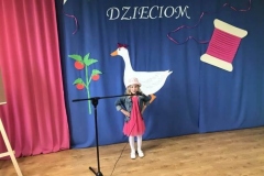 Zdjęcie przedstawia dziewczynkę ubraną w różową sukienkę,  białe rajstopy i biały kapelusz na głowie. Tło  stanowi niebieski materiał.