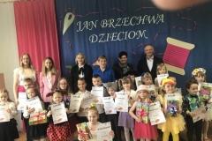 Grupa rodziców nauczycieli oraz dzieci bioracych udział w konkursie stoi na tle  niebieskiej dekoracji. Z boku wisi biało czerwona flaga. Dzieci trzymają w rękach dyplomy i upominki.