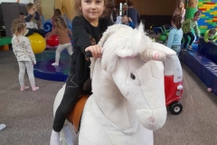 Dziewczynka siedzi i porusza sie na białym, dużym koniu. W tle widać inne dzieci.