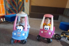 Dziewczynka siedzi w niebieskim autku, chłopiec w różowym  poruszają się nimi po dywanie.