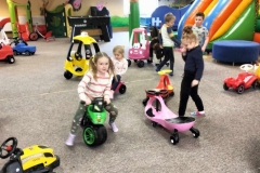 Dziewczynka siedzi na małym zielonym rowerku. Obok niej stoi druga.  W tle kolorowa  zjeżdżalnia.