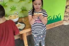 Dziewczynka stoi na tle zielonej ściany.  W rękach  trzyma kanapkę.