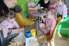 Przy dwóch stolikach siedzi czworo dzieci.  Na stoliku stoją dwa kubki i tosty.