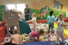 Trzy dziewczynki stoją wśród  kolorowych małych  kulek. Dwie  dziewczynki kucają. W tle kolorowa ściana.