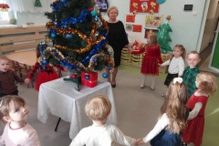 Na środku stoi kolorowa choinka. Wokół niej ustawione dzieci i pani. Trzymają się za ręce tworząc koło.