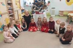 Grupa dzieci w odświętnych strojach siedzi w półkolu. Wśród nich pani w czarnej sukience. Za nimi na stoliku przykrytym białym obrusem stoi choinka.