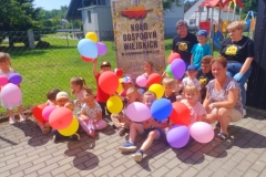 Dzieci siedzą na kostce, w rękach trzymają kolorowe balony, za nimi panie z Koła Gospodyń Wiejskich.