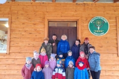 Grupa dzieci wraz z członkami Koła Łowieckiego stoją przed domkiem.