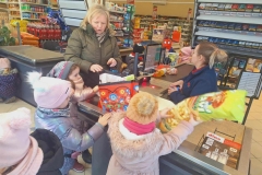 Dzieci stoją przy kasie. Pomagają pakowawać zakupy do dużej czerwonej torby. Jedna dziewczynka trzyma w rękach worek z chrupkami kukurydzianymi. Pani kasjerka w granatowym polarze kasuje zakupiony towar. Pani w zielonej kurtce wyciąga kartę płatniczą.