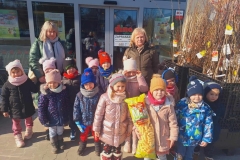 Grupa dzieci w kolorowych kurtkach, czapkach i szalikach stoi przed budynkiem. Za dziećmi 2 panie. Obok grupy z prawej strony pojemnik z sadzonkami drzew owocowych, z lewej paleta z workami ziemi. W tle duże, szklane drzwi.