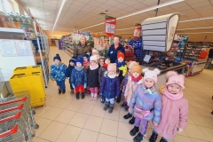 Grupa dzieci ustawiona w półkolu. Za nimi pani wychowawczyni i pani kasjerka. Obok drzwi ustawione żółte koszyki i srebrne na kółkach. W tle regały z napojami i słodyczami.