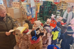 Pani w ciemnozielonej kurtce trzyma w rękach książkę. Obok niej stoi grupa dzieci. Za nimi pani w zielonej kurtce I popielatym szalu. Z boku pojemniki z papierem toaletowym.