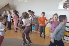 W sali przedszkolnej widoczna grupa dzieci będąca w ruchu - tańczą, podskakują. Ubrane są one w kolorowe spodnie i koszulki. W tle widać szafki przedszkolne. Na ścianie zawieszone jest godło. Po prawej stronie wisi szafka.