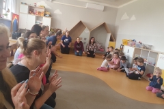 W sali przedszkolnej znajdują się dzieci i rodzice. Tworzą duży krąg, siedząc w siadzie klęcznym i po turecku.Mają uniesione w górę palce i pokazują zabawę paluszkową.. W tle szafki z zabawkami i pudełkami klocków.