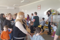 Grupa rodziców i dzieci bawią się w kole, tańczą do zabawy trzymając się za ręce.