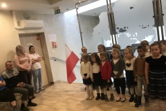 Dzieci ubrane w odświętne stroje stoją w dwuszeregu na tle szklanych elementów ściany. Pierwszy chłopiec stojący w tyle trzyma flagę.       Z lewej strony stoją dwie młode kobiety, za nimi są drzwi. Obok nich na krześle siedzi starszy mężczyzna oraz drugi częściowo tylko widoczny.