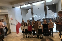 Dzieci ubrane w odświętne stroje stoją w dwuszeregu. Pierwszy chłopiec stojący  w tyle z lewej strony trzyma flagę, ten z prawej duże prostokątne godło. Z prawej strony stoi nauczycielka we wzorzystej bluzce i ciemnych spodniach. Jedna z dziewczynek ubrana w czarną sukienkę ze srebrnymi paskami stoi lekko wysunięta do przodu. Po lewej stronie widać stojące na tle drzwi trzy młode kobiety.