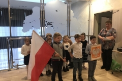 Na pierwszym planie trzej chłopcy. Pierwszy z lewej ubrany w szaro - ciemną bluzę i ciemne spodnie trzyma w dłoniach dużą flagę. Chłopiec    w środku ma białą koszulę oraz ciemne spodnie. Ten z prawej ubrany jest w białą koszulę i szarą marynarkę oraz szare spodnie. W dłoniach trzyma duże prostokątne godło. Z prawej strony stoi nauczycielka z białymi kartkami w dłoni. Ubrana jest we wzorzystą bluzkę i ciemne spodnie.