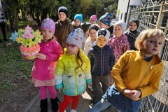 Grupa dzieci stoi ubrana w bramie przedszkola, dziewczynka trzyma w rękach koszyk z wiosennymi kwiatami.