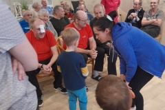 Chłopiec wita się z pensjonariuszem Domu Pomocy Społecznej na wózku inwalidzkim, w tle widać innych pensjonariuszy. Pani w niebieskiej bluzie pomaga chłopcu.