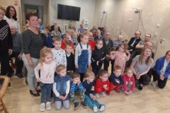 Kilkoro dzieci siedzi na podłodze, inne za nimi stoją, za dziećmi siedzą pensjonariusze Domu Pomocy Społecznej.