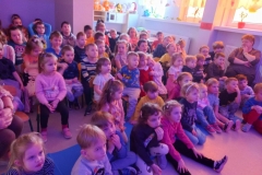 Grupa dzieci w kolorowych strojach siedzi na podłodze. Pozostałe siedzą na krzesełkach. Za nimi widać okna.