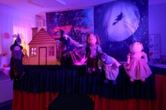 Zdjęcie przedstawia cztery lalki siedzące na scenie. Obok nich jest niewielki drewniany domek.