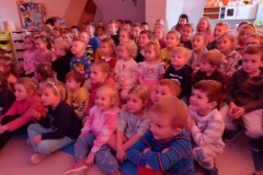 Grupa dzieci siedzi na podłodze, pozostałe siedzą na krzesłach.