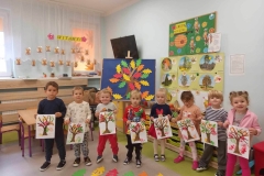 Ośmioro dzieci  stoi w półkolu. Każde trzyma przed sobą kartkę,  a na niej drzewo z kolorowymi liśćmi.  W tle obrazki z drzewami.