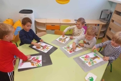 Pięciu chłopców siedzi przy  stoliku i stemplują kolorowe liście na brązowych drzewach. W tle jasna ściana.