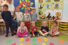 Czworo dzieci siedzi na podłodze, za nimi stoi kolejna czwórka. Na podłodze widnieje napis dzień drzewa napisany na kolorowych liściach. W tle na niebieskim płótnie widać kolorowe liście. Obok tablica, a na niej zielone drzewa.