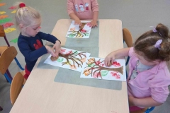 Trzy dziewczynki siedzą przy  stoliku, przed nimi białe kartki, a na nich kolorowe liście.