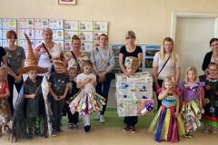 Grupa dzieci stoi w kolorowych strojach wykonanych z różnych materiałów przeznaczonych do recyklingu, za nimi stoją rodzice.