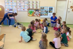 Dzieci zgromadzone wokół stołu, wykonują eksperyment. Wkładają do słoiczkow różne rzeczy, zakręcają i zakopują do ziemi.