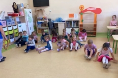 Dzieci siedzą w kole przed tablicą, słuchają czytanego wiersza, rodzic układa śmieci występujące w wierszu.