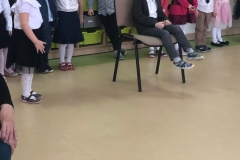 Na krześle siedzi dziadziu-chłopiec, dziewczynka zadaje mu pytanie, chłopiec odpowiada. W tle za chłopcem widać dzieci ustawione w rzędzie.