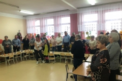 Na zdjęciu widać babcie i dziadków. Stoją przed stolikami. Dzieci śpiewają im sto lat.