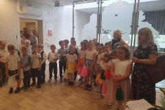 Grupa dzieci ustawiona w półkolu. W rękach trzymają kolorowe pompony. Przy nich stoją 2 panie w ciemnych sukienkach.