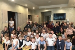 Grupa dzieci w eleganckich strojach i 3 panie siedzą na podłodze. Za nimi na krzesłach siedzą staruszkowie- mieszkańcy DPS- u. W drzwiach stoi pani w białym fartuchu i niebieskich spodniach.