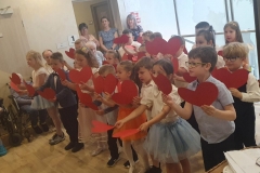 Dzieci w kolorowych strojach ustawione są w 3 rzędach. W obydwu rękach trzymają czerwone serduszka. Za nimi stoją 3 panie.