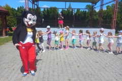 Zdjęcie przedstawia postać z bajki - myszkę. Ubraną w czerwone spodnie i biało czerwony frak. Za nią dzieci w rzędzie.