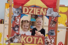 Dwie dziewczynki stoją za przedszkolnym teatrzykiem w prostokątnej brązowej ramce. Brzegi ramki ozdobione zdjęciami kotów z literami układającymi się w napis Dzień Kota. W tle przedszkolna szafka z zabawkami i książeczkami dla dzieci.